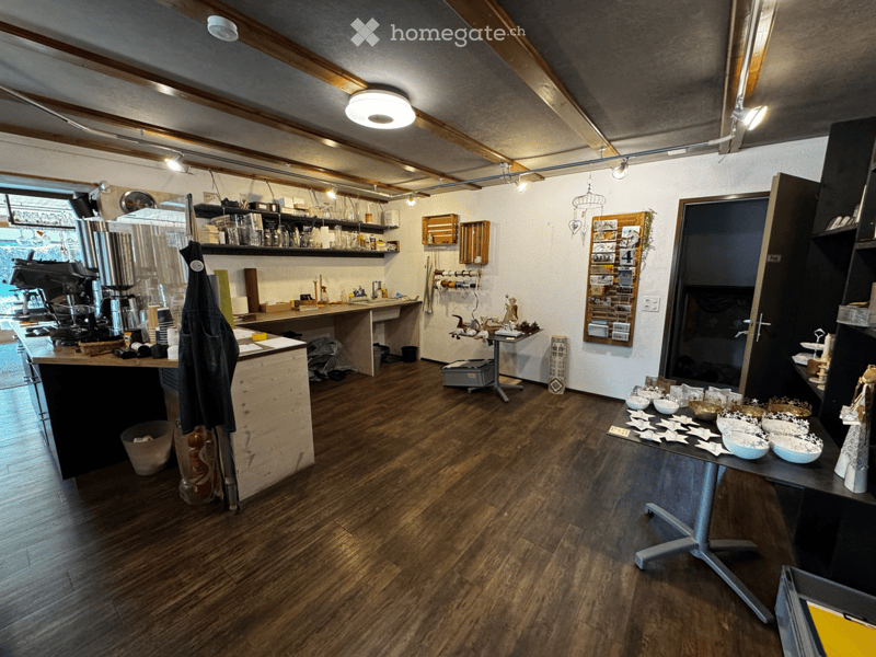 A modern, well-lit room with wooden flooring, exposed wooden beams, and a coffee machine on the counter. Shelves hold cups and other items, and a small table displays decorative bowls and star-shaped items.