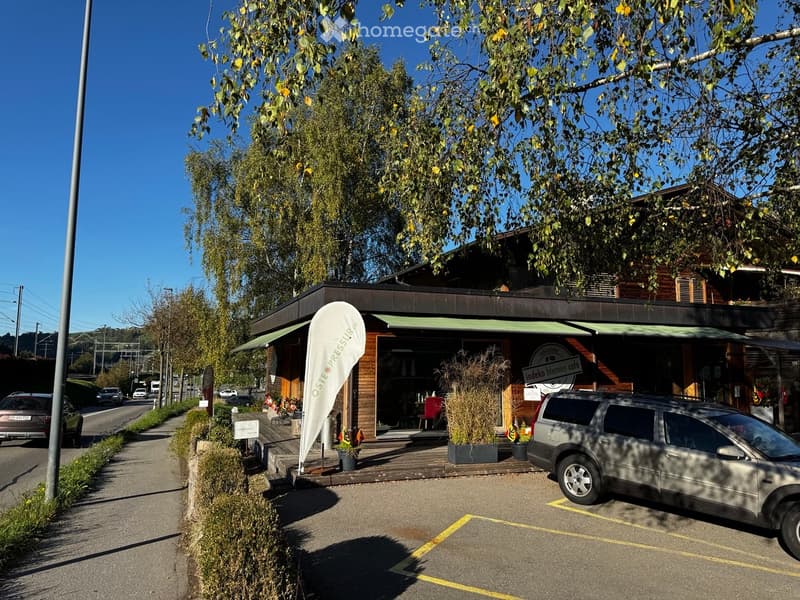 A modern wooden cafe building with a green awning, outdoor seating area, and a parked car in front.