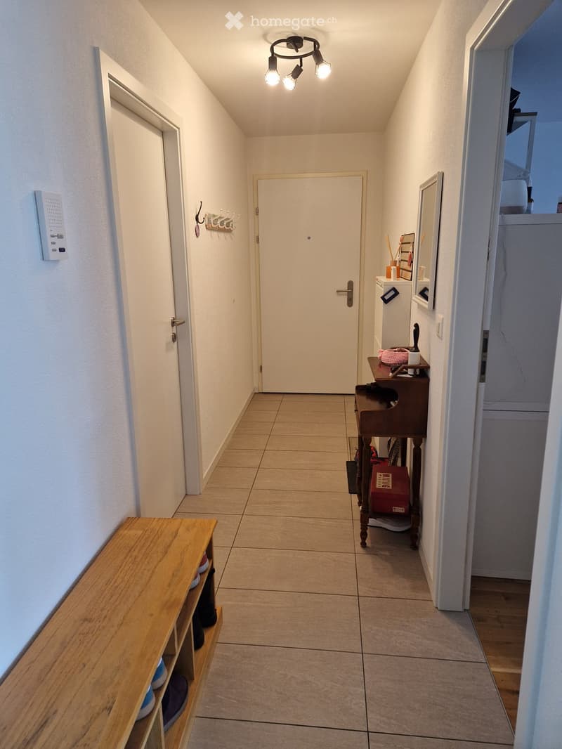 A narrow hallway with tiled flooring, white walls, and a ceiling light fixture. There are two wooden benches with shoes and bags, a small wooden table with items, and a coat rack with hooks.
