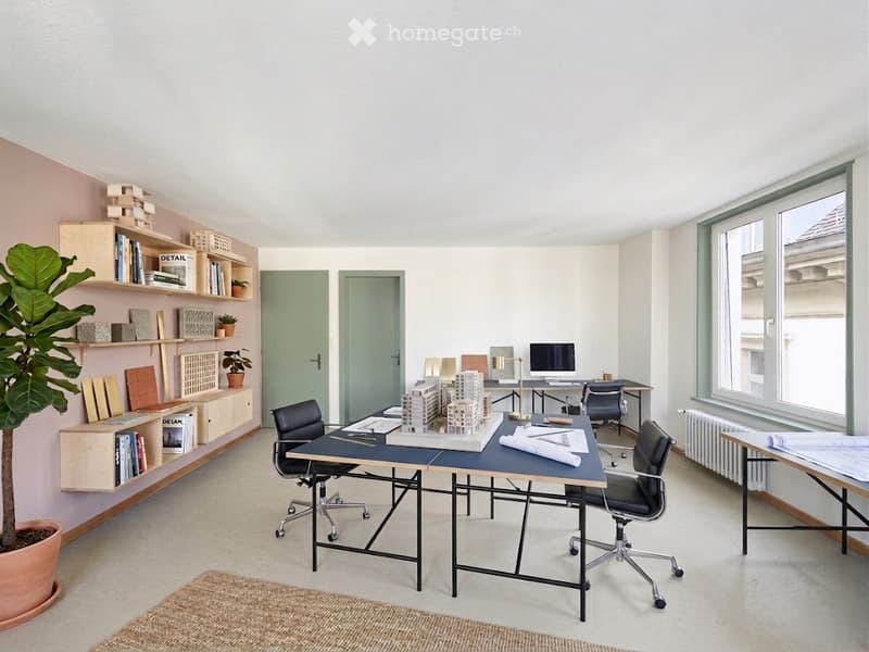 A modern, well-lit office space with a large central table, chairs, shelves filled with books and design samples, a computer, and a potted plant. The room features large windows and a minimalist aesthetic.