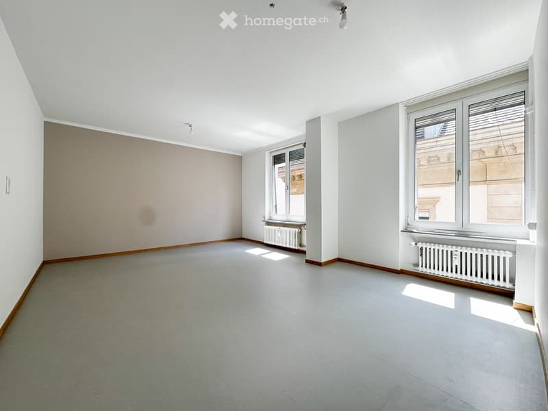 Empty room with light grey walls, wooden baseboards, grey carpeted floor, and two windows with radiators beneath them.