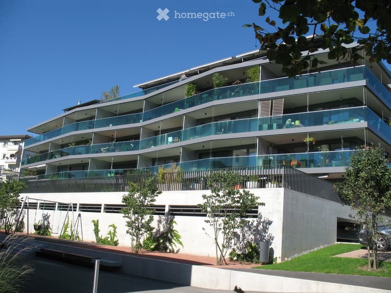 Modern multi-story building, glass balconies, concrete base, plants and trees