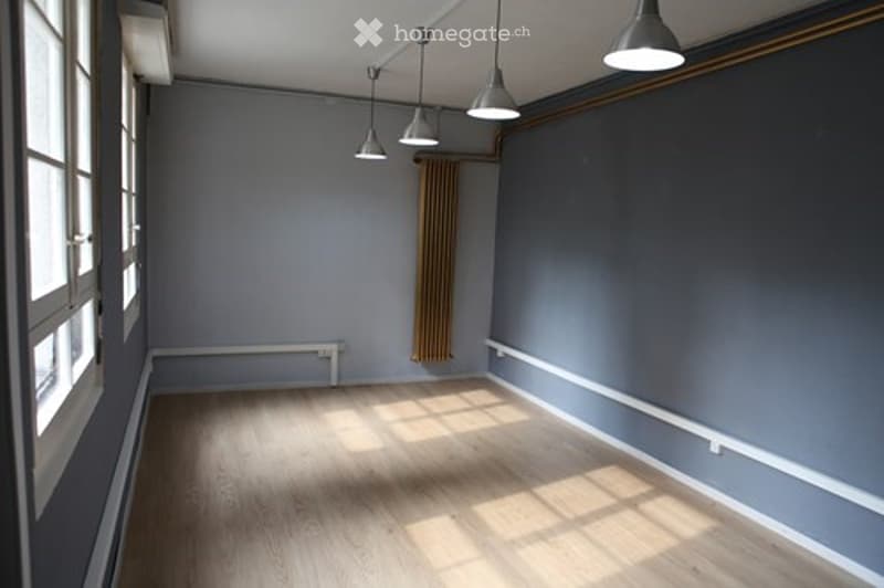 Empty room with wooden flooring, grey walls, large windows, and pendant lights.