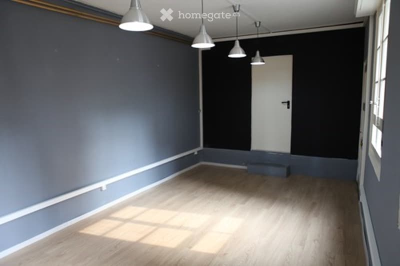 Empty room with grey walls, light wooden flooring, white door, and pendant lights.