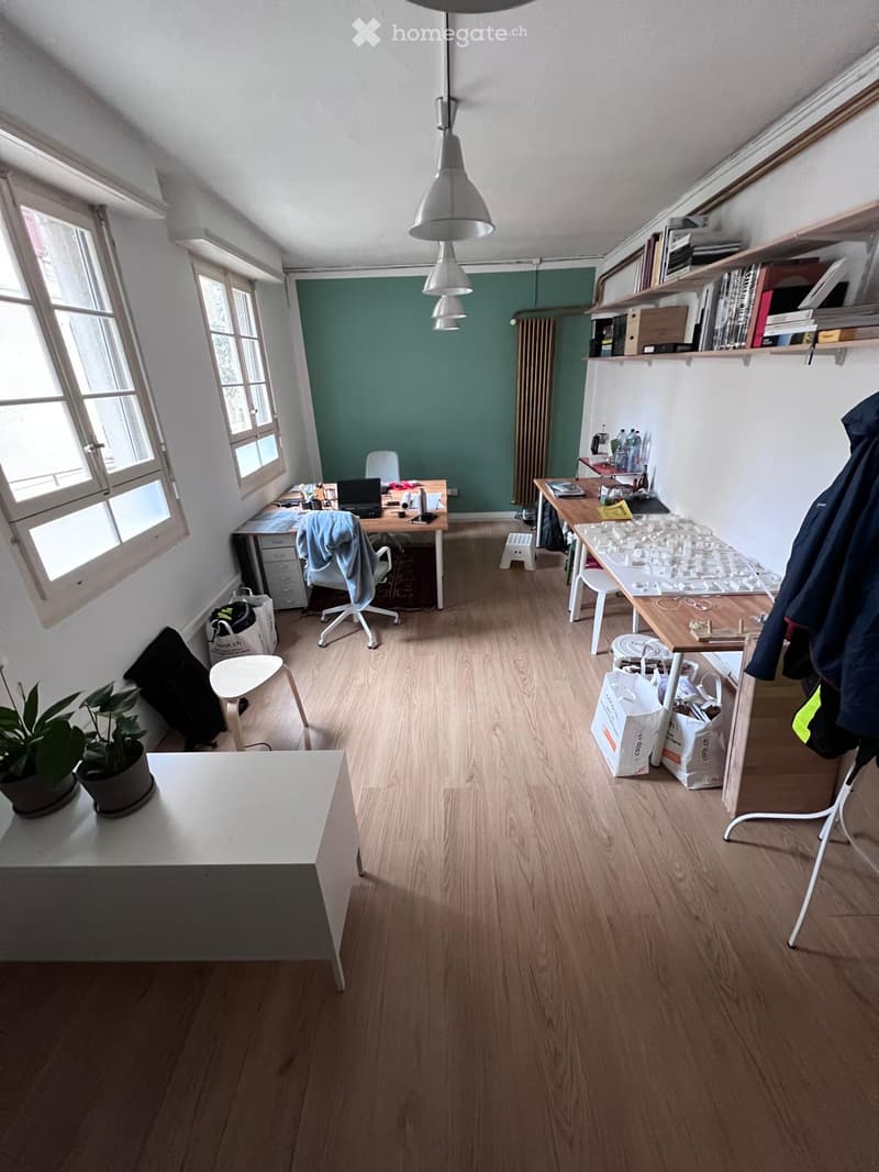 Bright, open room with wooden flooring, multiple desks, chairs, shelves, and a potted plant. Natural light from large windows illuminates the space.