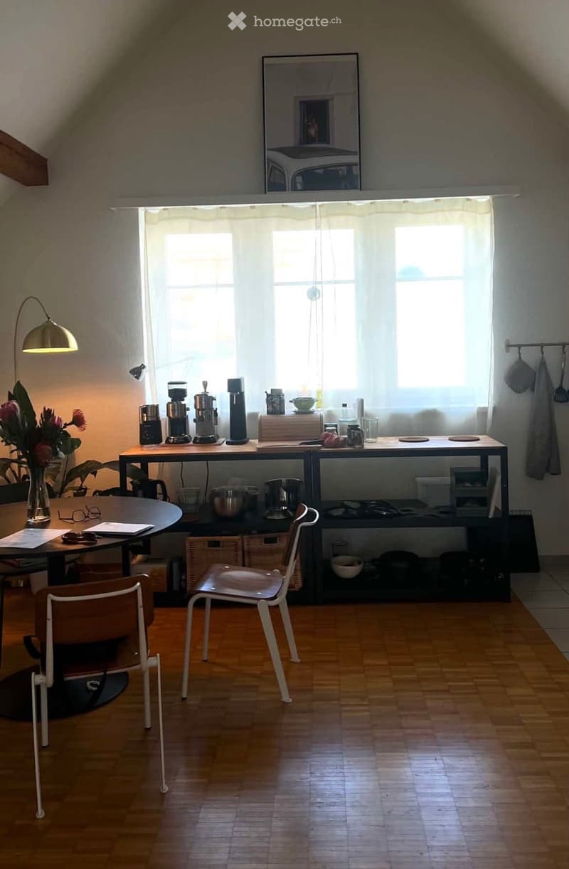 Bright kitchen with wooden flooring, large windows, coffee maker, table, chairs, and decorative vase with flowers.