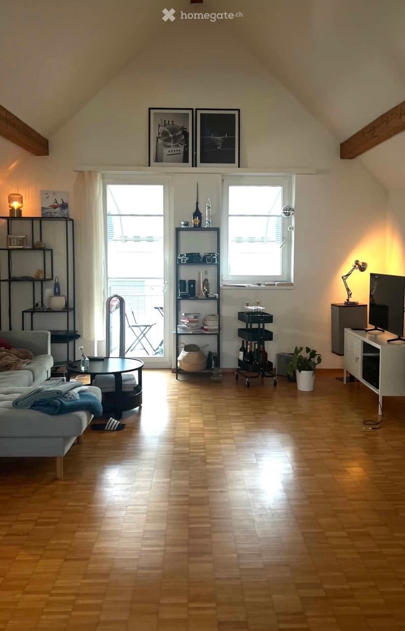Bright living room with wooden flooring, large windows, and a sliding glass door leading to a balcony. Features a grey sofa, coffee table, shelves with books and decor, and a TV stand with a television.
