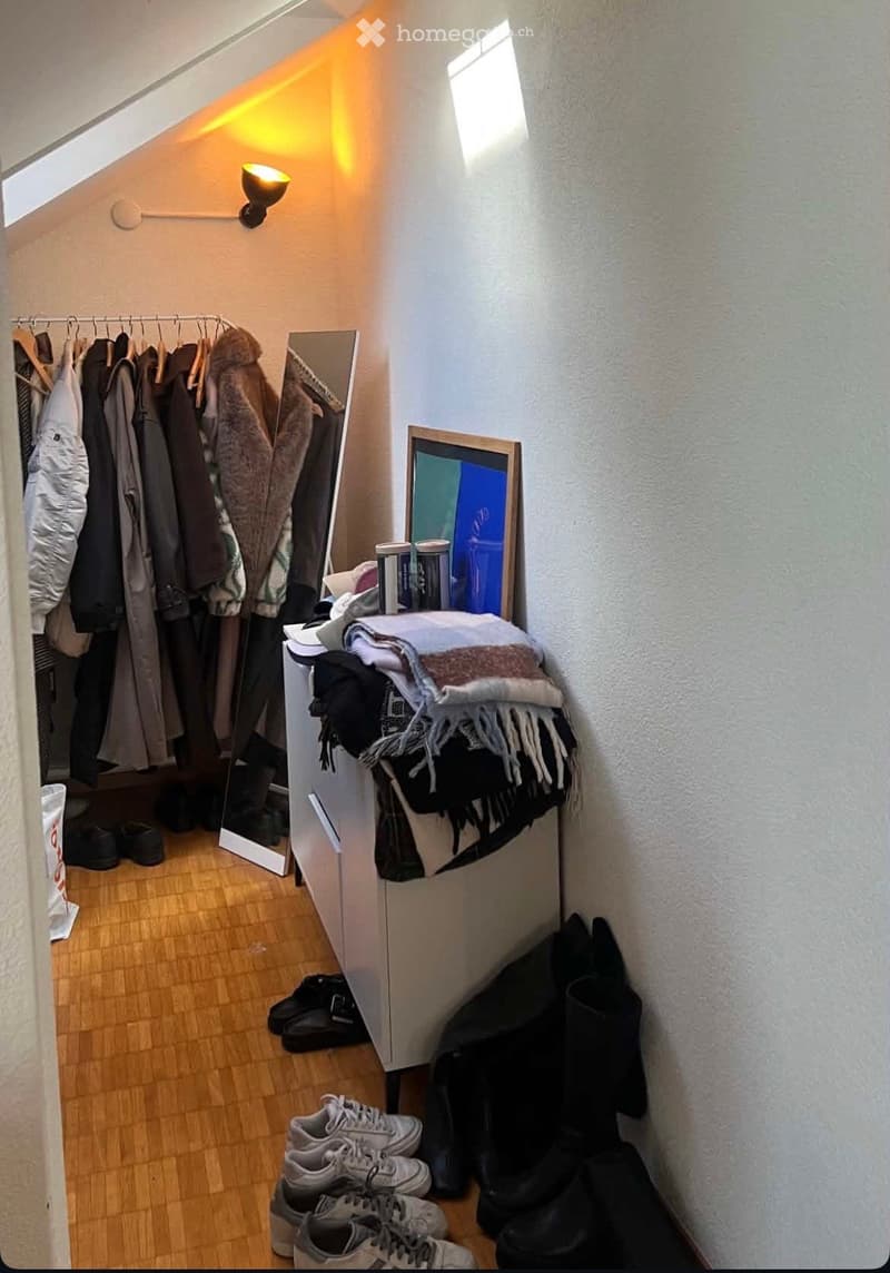 Attic room with wooden flooring, slanted ceiling, and large window. Contains a wardrobe with hanging clothes, a mirror, a picture frame, books, folded blankets, and several pairs of shoes.