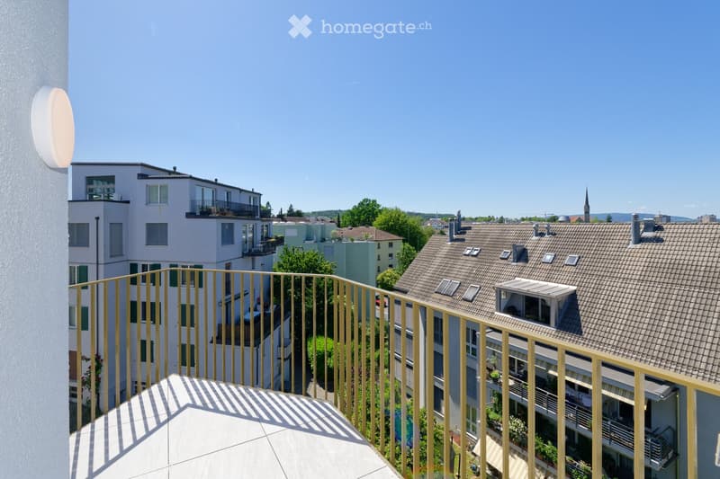 Balcony overlooking a residential area with multiple buildings, green trees, and a clear blue sky.