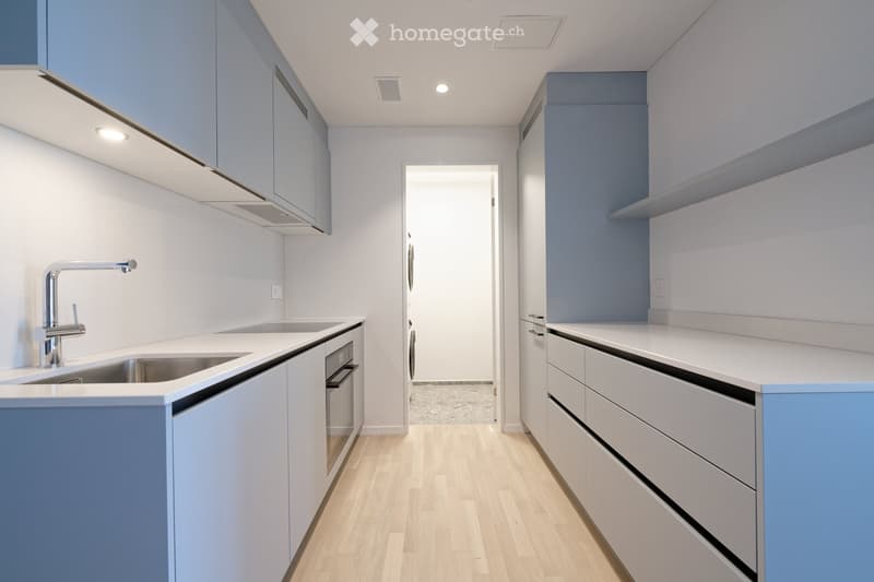 Modern kitchen with white countertops, light blue cabinets, and a stainless steel sink with a contemporary faucet. The space features wooden flooring and built-in storage.