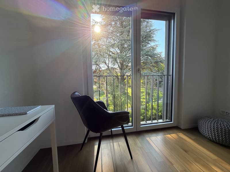 Bright room with wooden flooring, a modern chair, a small table with a book, and a round stool near a large balcony door with a view of trees.