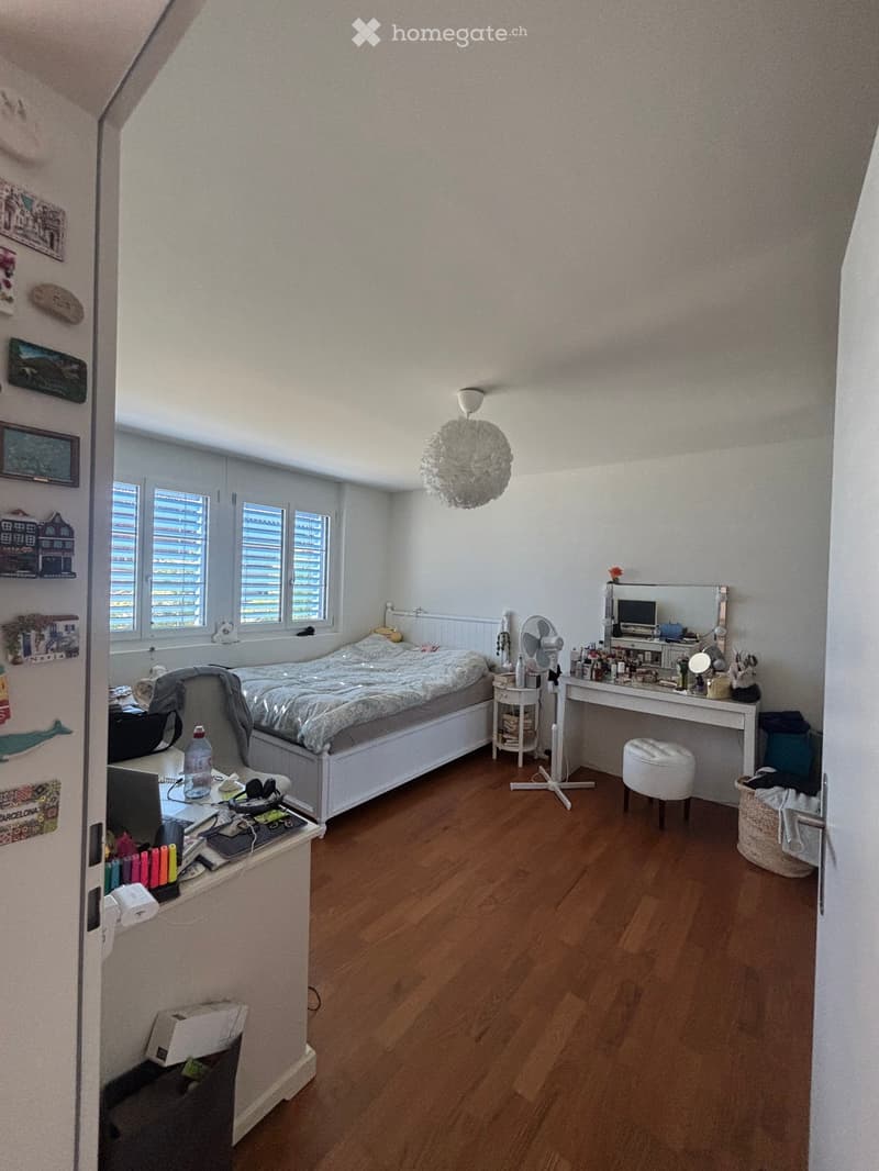 Well-lit bedroom with wooden flooring, a bed with a patterned duvet, a desk with a laptop, a vanity mirror, and a small fan. The room features large windows with blinds and a hanging decorative light fixture.