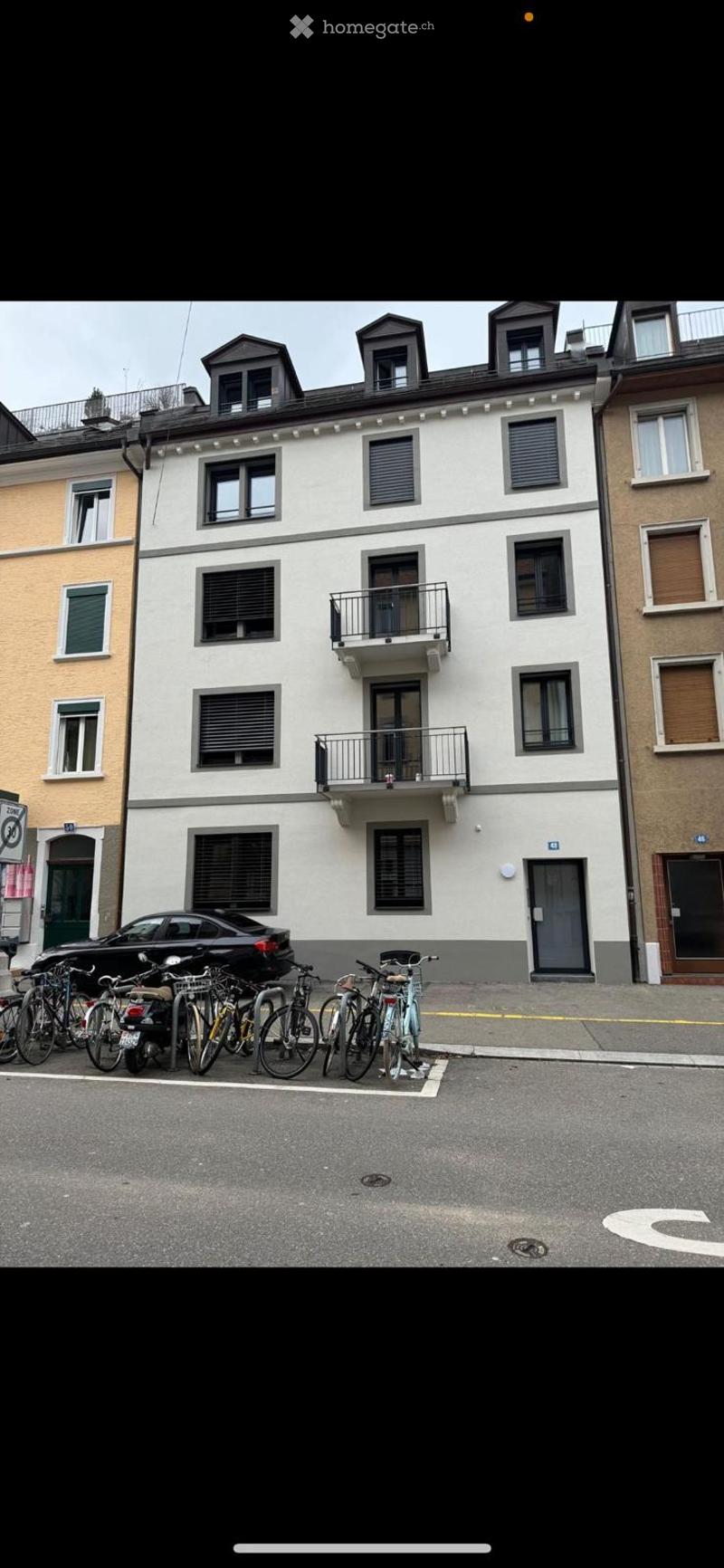 A multi-story residential building with white facade, multiple windows, and balconies. Several bicycles and a car are parked in front.