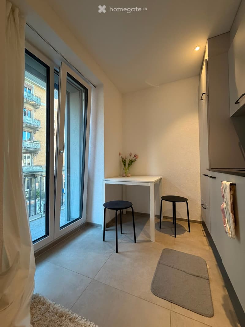 Small, well-lit room with a white table, two black stools, a vase with flowers, a gray rug, and a curtain covering a large window that offers a view of a balcony.
