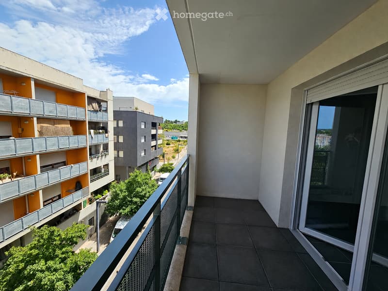 Balcony with tiled floor, metal railing, and sliding glass door leading inside. View includes neighboring apartment buildings, greenery, and a clear blue sky.
