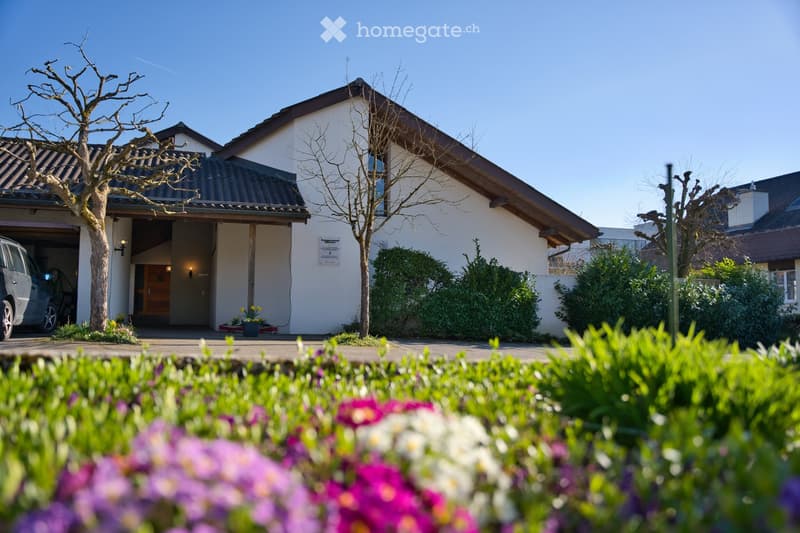 Two-story house with a garage, white exterior, and a well-maintained garden with colorful flowers in the foreground.