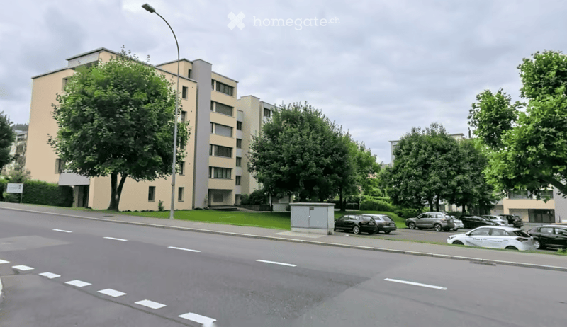 Multi-story residential building with light-colored facade, multiple windows, and surrounding greenery. Several parked cars are visible along the street.