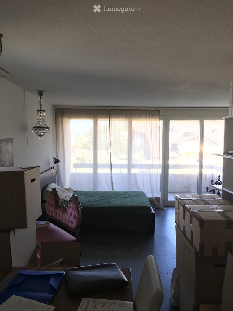 A bedroom with a bed, a chair, a table, and a large window with curtains. The room has a chandelier and a lamp on the wall.