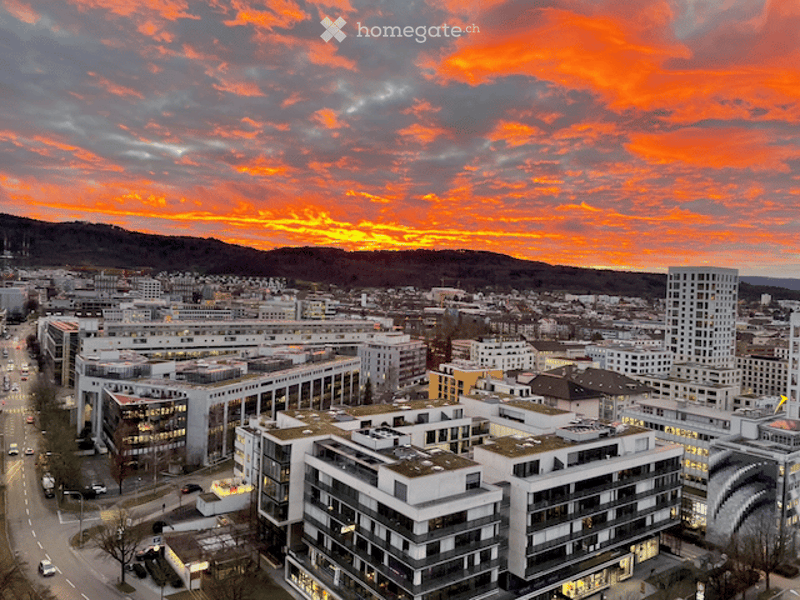 Aerial view of a cityscape at sunset, featuring modern buildings, roads, and a distant mountain range.