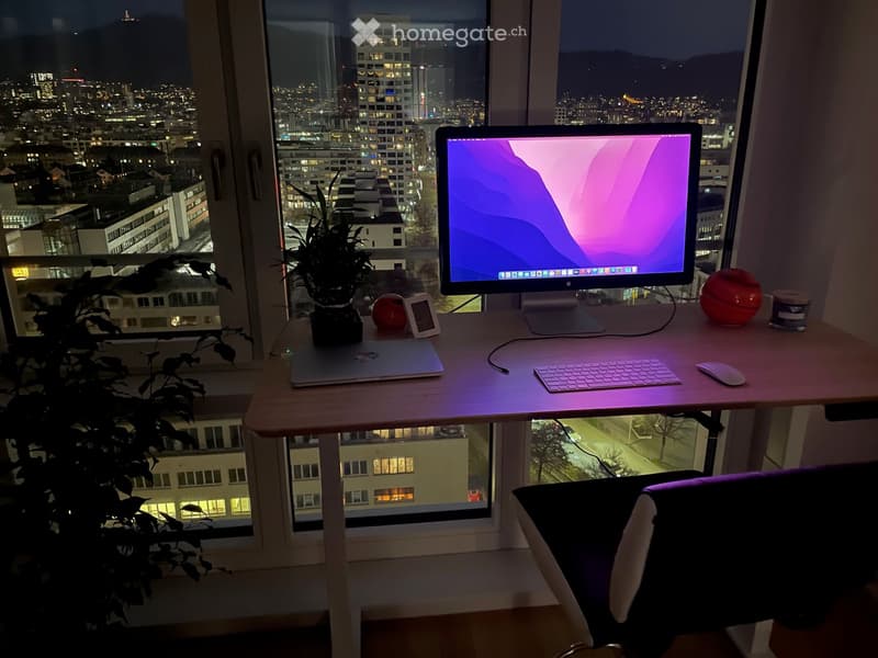 A modern workspace with a desk, laptop, monitor, keyboard, mouse, chair, plant, and decorative items, overlooking a cityscape at night.