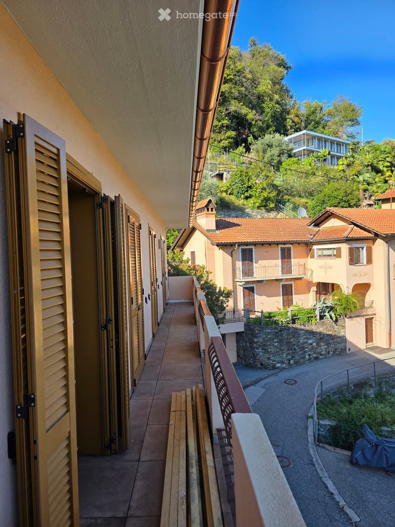 Balcony with wooden bench and railing, overlooking a narrow street lined with houses and greenery.
