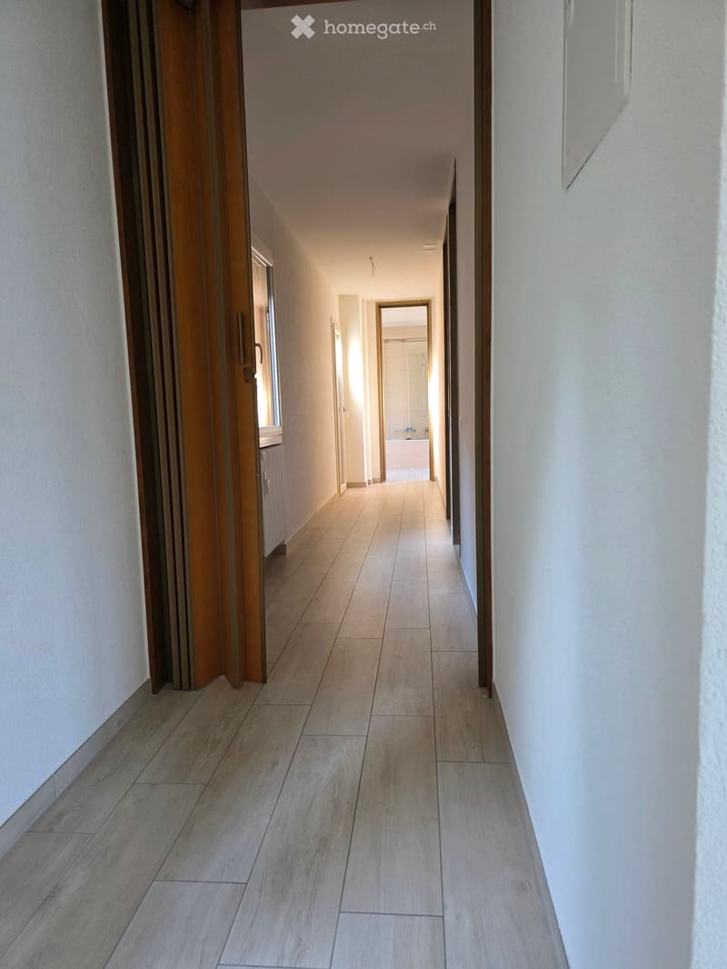 A long, narrow hallway with light-colored tiled flooring and white walls. Doors on both sides lead to other rooms, with a visible bathroom at the end.