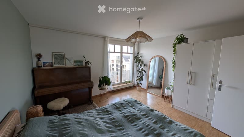 Cozy bedroom with a bed, piano, white wardrobe, large mirror, and several potted plants. The room features wooden flooring and large windows with a view of the city.
