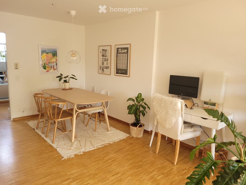 A modern, well-lit room with a wooden table, chairs, a white desk with a screen, and decorative plants.