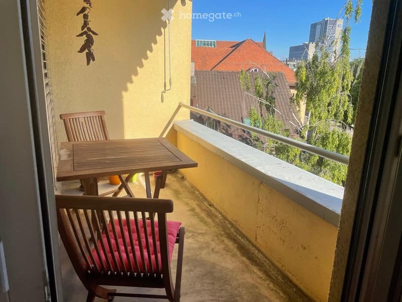 Balcony with wooden table and chairs, overlooking a cityscape with buildings and trees.