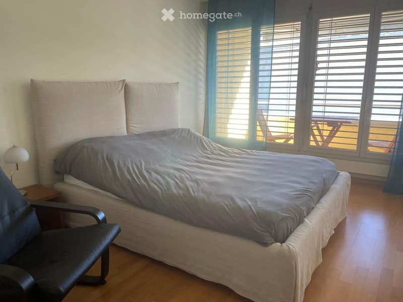 Cozy bedroom with a large bed, nightstand, table lamp, and a black leather chair. Large windows with blinds lead to a balcony.