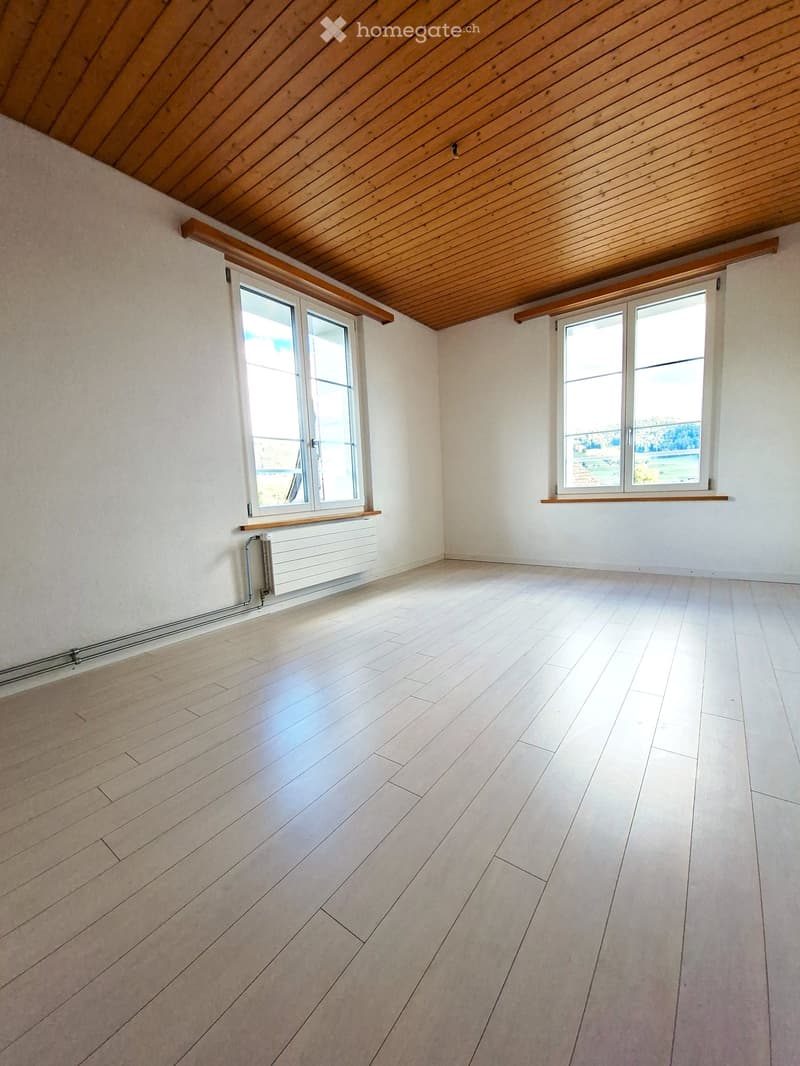 Empty room with light wooden flooring, white walls, and large windows offering a view. The ceiling is wooden with exposed beams.