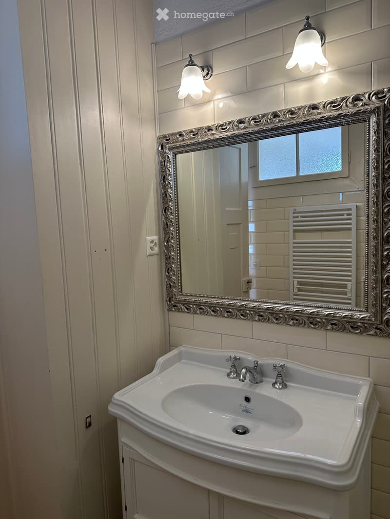 Bathroom with a white sink, ornate silver-framed mirror, and a radiator. The walls are tiled in light beige, and the sink has a modern chrome faucet.