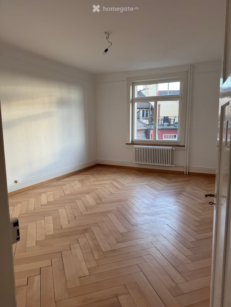 Empty room with light wooden herringbone flooring, white walls, a single window with radiator below, and a visible doorway.