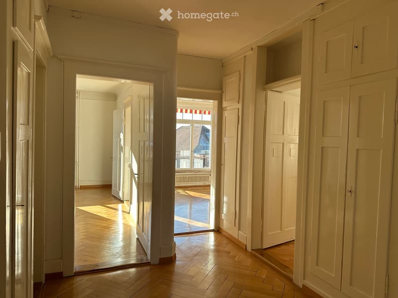 Empty hallway with wooden flooring, white walls, multiple doors, and a view of a room with large windows and a radiator.