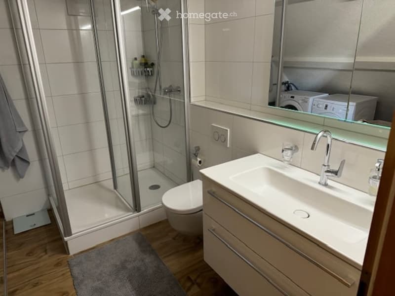 Modern bathroom with a white tiled shower, a white sink with a chrome faucet, a toilet, and a large mirror above the sink. A grey towel hangs on the wall, and a bath mat is on the floor.