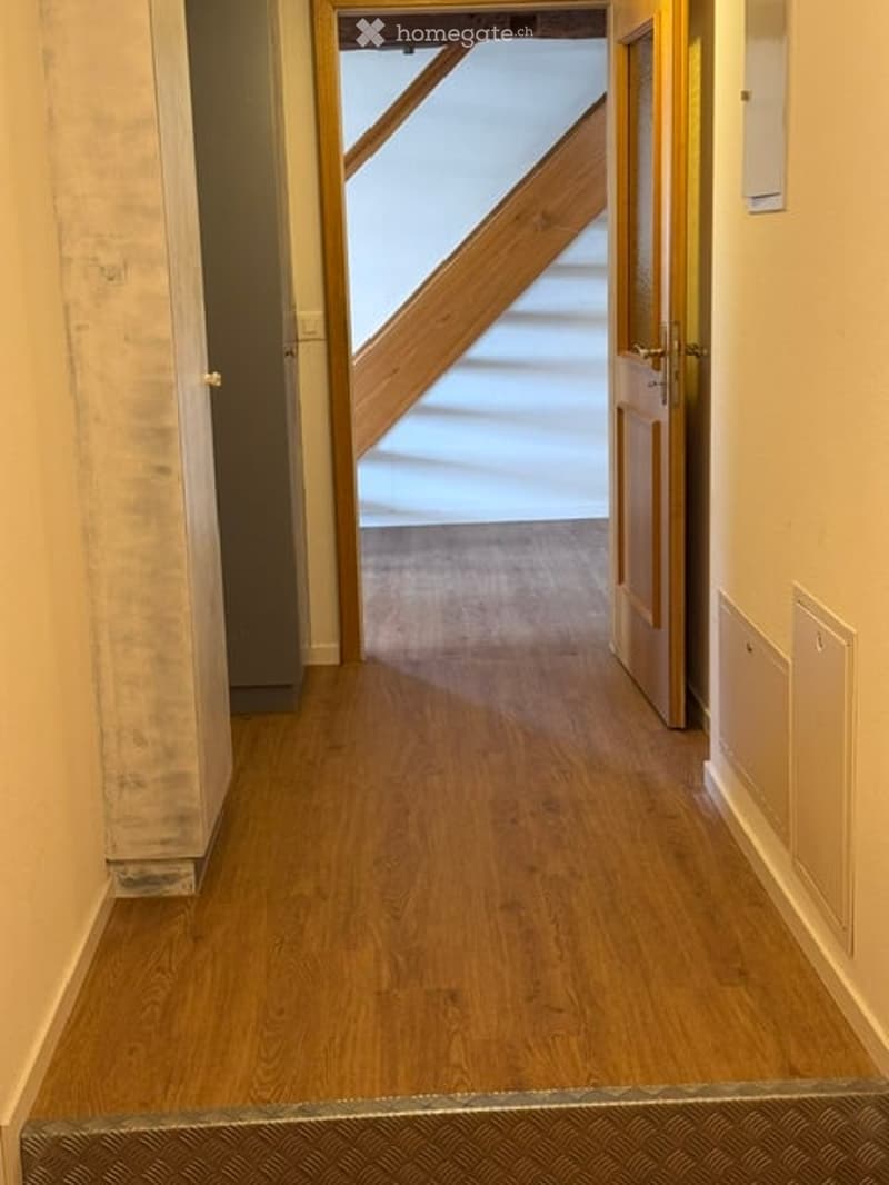 A narrow hallway with wooden flooring, white walls, and a staircase visible at the end.