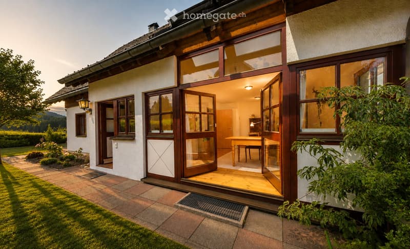 A modern two-story house with large glass doors leading to a wooden deck, surrounded by a well-maintained garden and lawn.