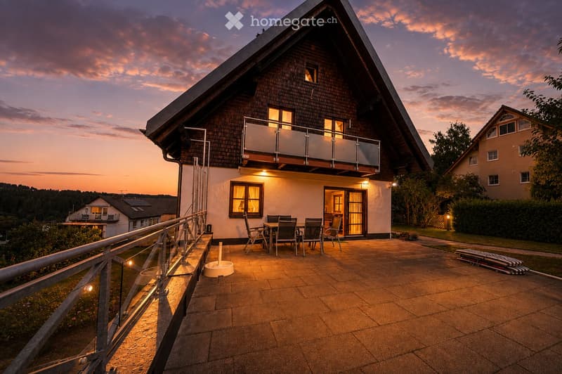 Balcony with a table and chairs, overlooking a house with a modern design and wooden exterior.