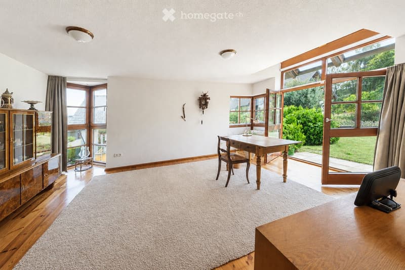 Brightly lit room with wooden flooring, large windows, and a view of greenery. Features a wooden chair, a table with a lamp, a cabinet with glass doors, and a large area rug.