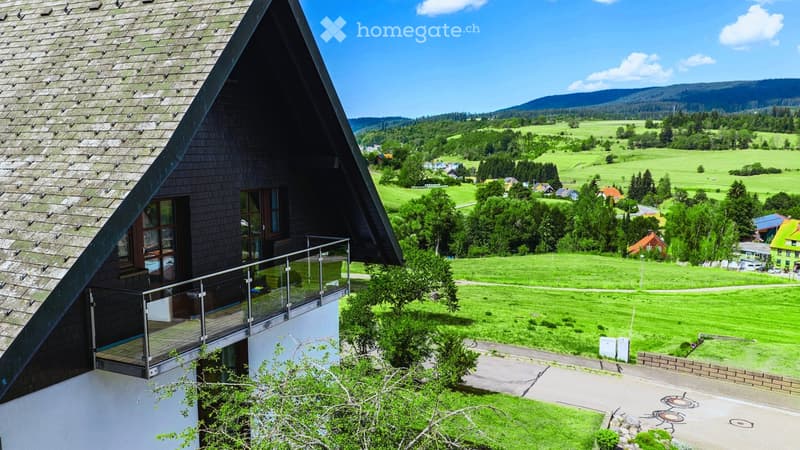 Modern house with a triangular roof and large windows, overlooking a scenic valley with green fields and scattered houses.