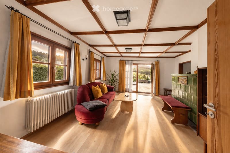 Bright living room with wooden beams, large windows, red sofa, coffee table, bench, radiator, and fireplace.
