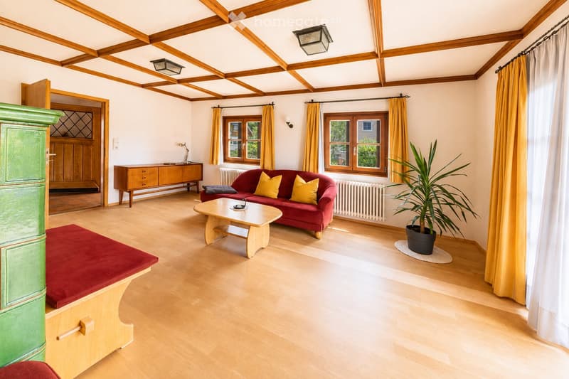 Bright living room with wooden floors, exposed wooden ceiling beams, large windows with yellow curtains, red sofa with yellow cushions, coffee table, sideboard, and a potted plant.