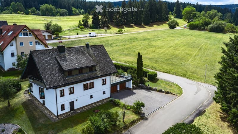 Aerial view of a two-story house with a shingled roof, white exterior, and a small balcony. The house is surrounded by a well-maintained lawn, trees, and a winding driveway leading to a garage.