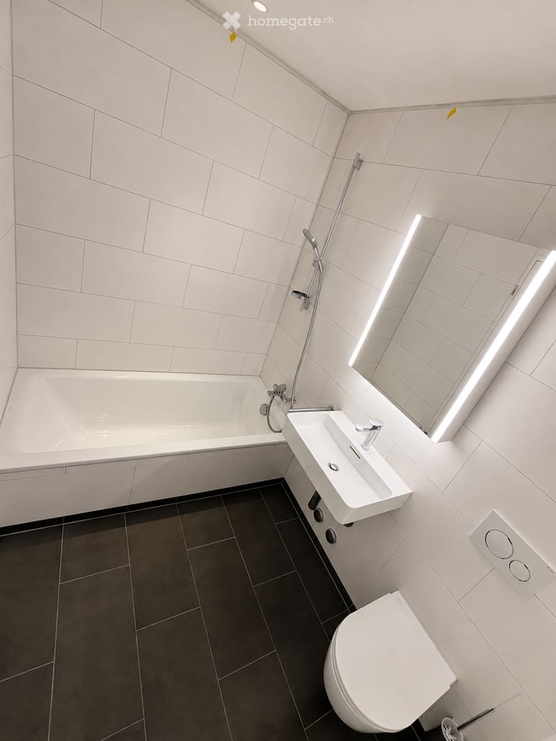 Modern bathroom with white tiled walls, a bathtub, a sink, a toilet, and a large mirror with built-in lighting.