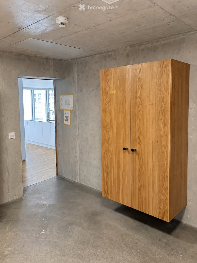 Empty room with exposed concrete walls and floor, a single wooden cabinet, and a doorway leading to another room with a visible kitchen area.