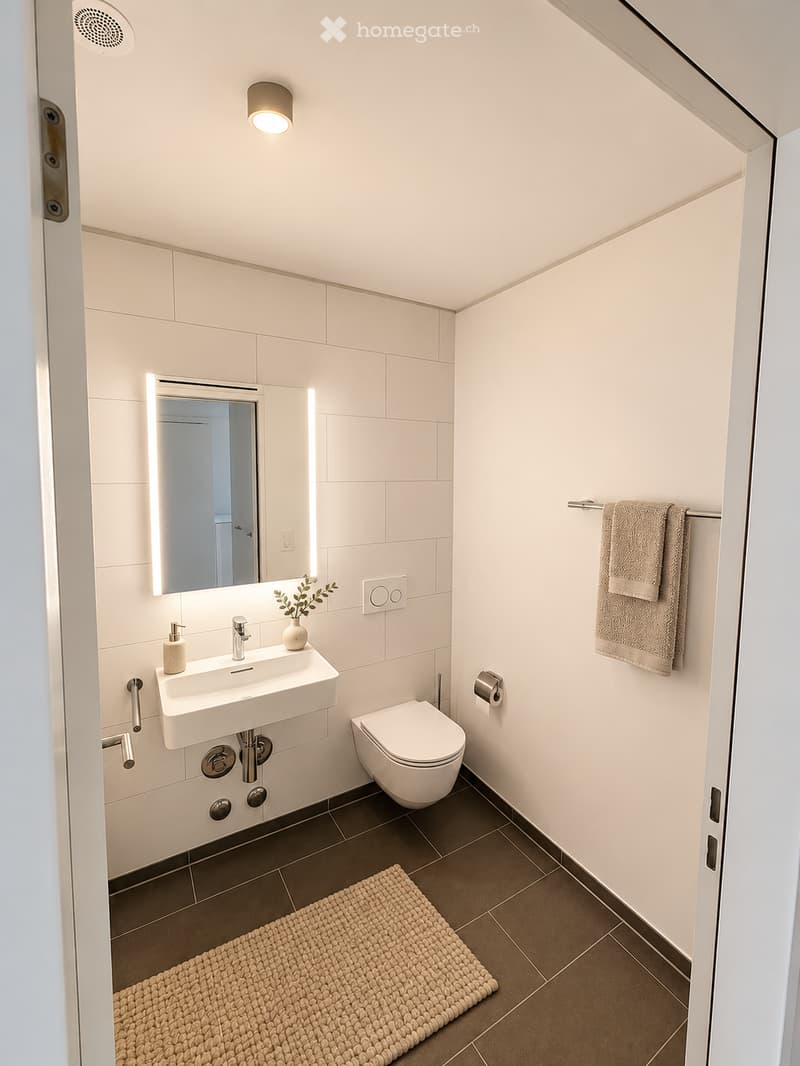 Modern bathroom with white tiles, a floating sink, a toilet, a mirror with built-in lighting, a towel rack, and a small decorative plant on the sink.