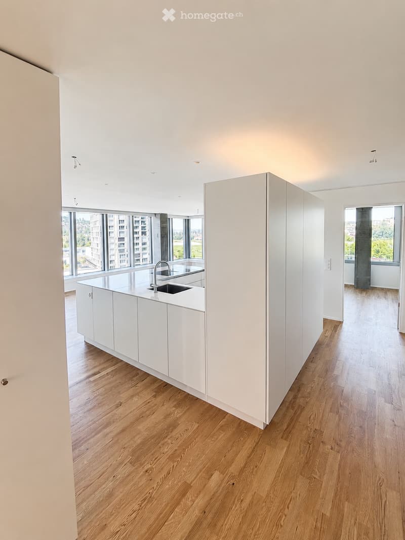 Modern, minimalist kitchen with white cabinetry, a central island, and a sink, featuring large windows and wooden flooring.