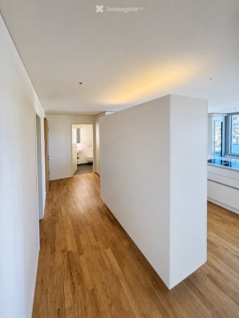 Bright, minimalist hallway with light wooden flooring and white walls. Features recessed ceiling lighting and open doorways leading to adjacent rooms, including a bathroom and a room with a window and built-in cabinetry.