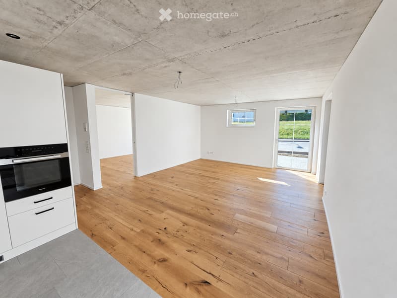 Empty room with light wooden flooring, white walls, and a modern concrete ceiling. Features include a built-in oven, a large window, and a sliding glass door leading outside.