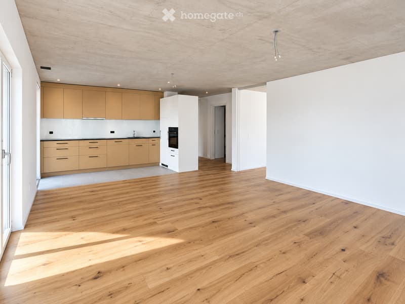 Open-plan kitchen and living area with light wooden flooring, modern light wood cabinets, and a built-in oven. The space is bright with large windows and recessed lighting.